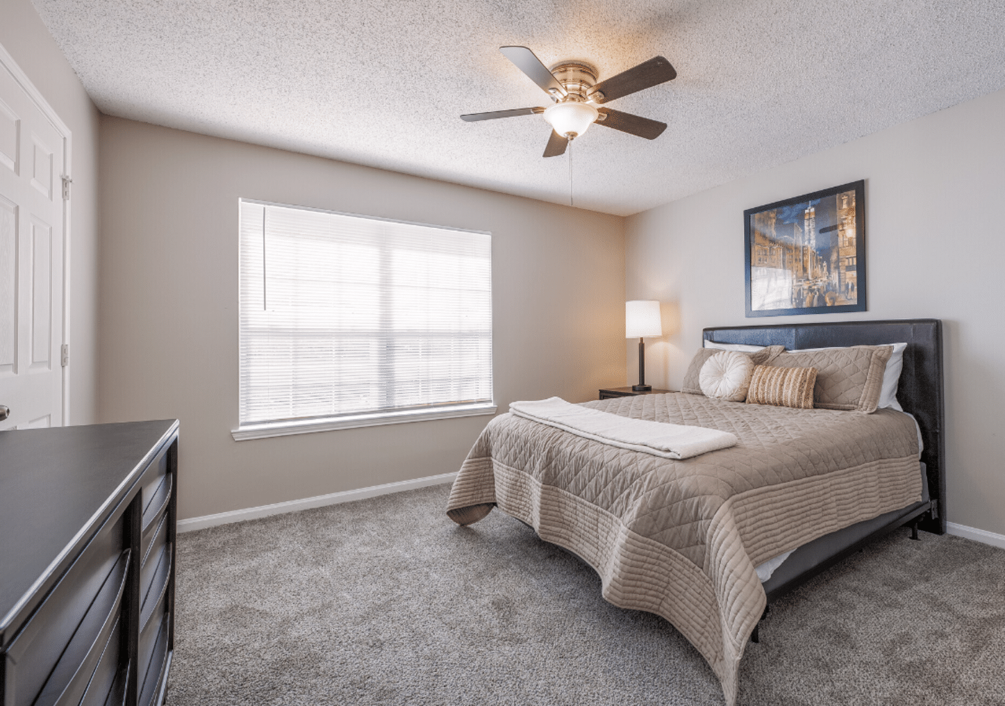 A neatly made bed with beige bedding in a carpeted bedroom, featuring a ceiling fan, dresser, lamp, and framed artwork on the wall. Natural light enters through a window.