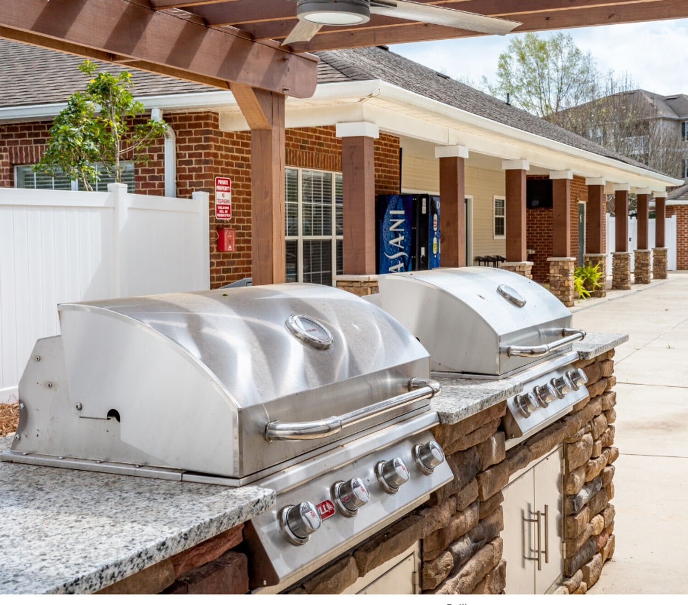 Two stainless steel outdoor grills on a stone and granite countertop under a pergola, with brick buildings and a vending machine in the background.